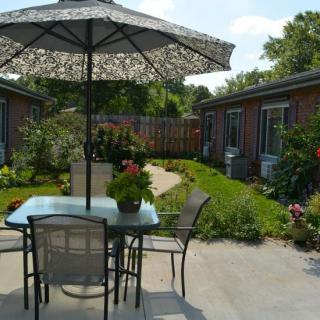 A serene patio setting with a table and chairs under an umbrella, surrounded by a lush garden with a winding pathway.