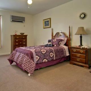 A cozy bedroom with a single bed adorned with purple bedding, featuring a nightstand with a lamp, an air conditioner on the wall, and a wooden dresser.
