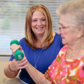A healthcare professional assists a senior woman with exercise using a dumbbell.