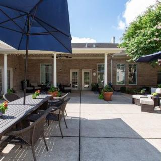 A sunny outdoor patio with seating, umbrellas, and potted plants in front of a brick building.