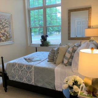 A cozy, well-decorated bedroom featuring a bed with multiple pillows, a lamp on the side table, artwork on the wall, and a window providing natural light.