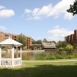 A scenic view of a park with a gazebo, lake, and surrounding apartment buildings under a partly cloudy sky.