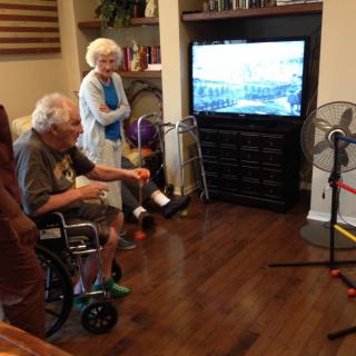 An elderly man in a wheelchair and an elderly woman are watching TV in a cozy living room.