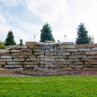 A small waterfall cascading over a stone wall in a landscaped garden setting.