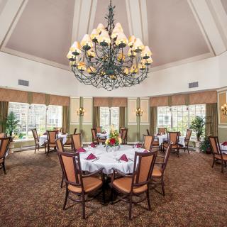 An elegant dining room with a large chandelier, neatly arranged tables and chairs, and large windows offering natural light.
