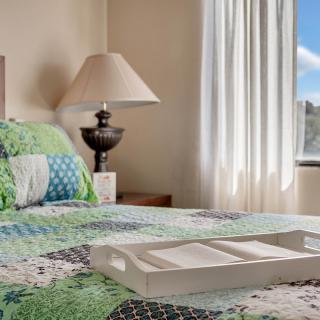 A cozy bedroom with a quilted bedspread and a decorative lamp, illuminated by natural light from the window.