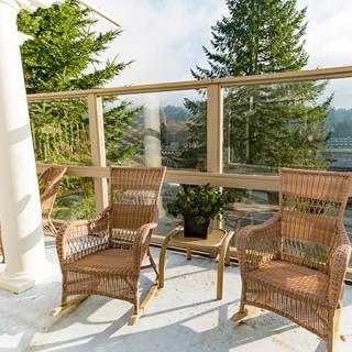 A peaceful patio setup with wicker chairs, surrounded by glass railings and greenery, on a sunny day.