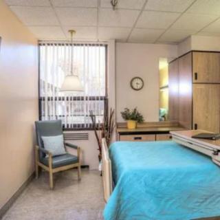 A tidy hospital room with a bed, chair, and window featuring vertical blinds.
