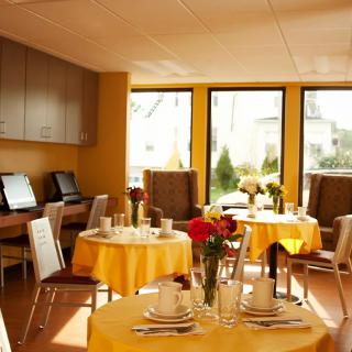 A cozy dining area with tables set for guests, featuring bright yellow tablecloths and natural lighting from large windows.