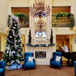 A festive lounge area featuring a large Christmas tree, elegant chandelier, and cozy blue chairs arranged around a fireplace.