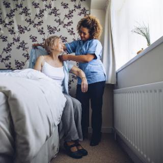 A caregiver assists an elderly patient in a cozy bedroom setting.