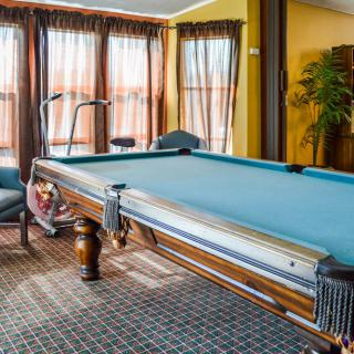 A cozy game room featuring a classic pool table, surrounded by large windows with drapes, and comfortable seating on a patterned carpet.