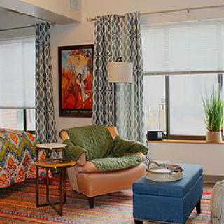 A modern living room with colorful decor, featuring a comfortable armchair, a blue ottoman, patterned curtains, and vibrant artwork.