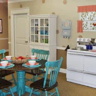 A cozy kitchen and dining area with vintage decor, featuring turquoise chairs around a round table and a white cabinet.