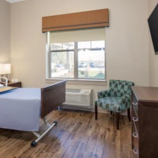 A hospital room with a bed, chair, and television, featuring wooden flooring and a window with natural light.
