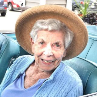 An elderly woman in a hat, smiling while seated outdoors.
