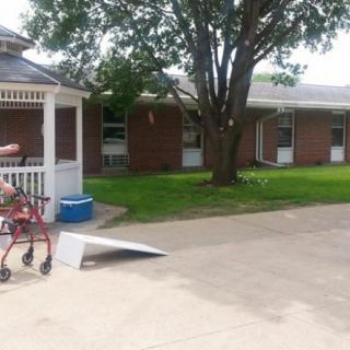An elderly person walking outdoors with a mobility aid near a house, with a tree and wheelchair ramp visible.