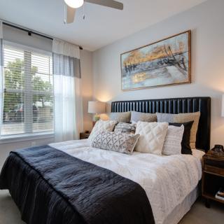 A modern bedroom featuring a large bed with decorative pillows, a stylish headboard, and wall art. Natural light streams in through the window, complementing the cozy ambiance created by the bedside lamp.