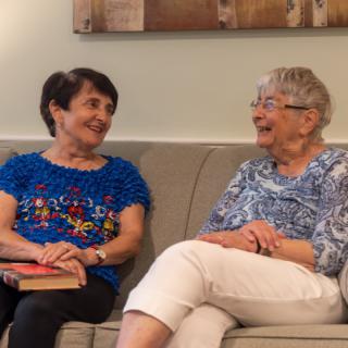 Two elderly women sitting on a couch, smiling and talking to each other.