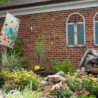 A vibrant garden with colorful flowers and yard decor in front of a brick wall adorned with window shutters.