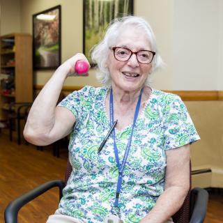 An elderly woman smiling while exercising with a pink ball indoors.