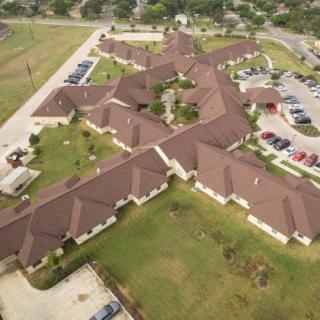 Aerial view of a large building complex with a distinctive roof structure surrounded by green spaces and parking lots.