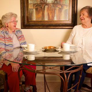 Two elderly women enjoying coffee and conversation at a glass table indoors.