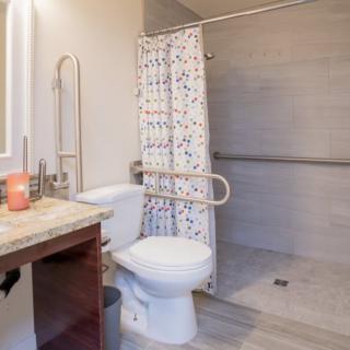An accessible bathroom featuring a toilet with grab bars, a shower area with a curtain, and a sink with a mirror.