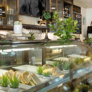 A cozy cafe with a variety of sandwiches displayed in a deli counter, adorned with small potted plants and a menu board in the background.