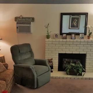 A cozy living room with a recliner facing a tiled fireplace, adorned with framed pictures and flanked by a lamp and indoor plants.