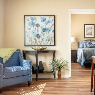 A cozy living room with a blue chair and artwork, leading into a bedroom with soft lighting.