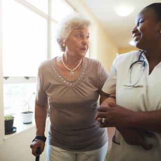 A nurse assists an elderly woman with a walking aid down a hallway.