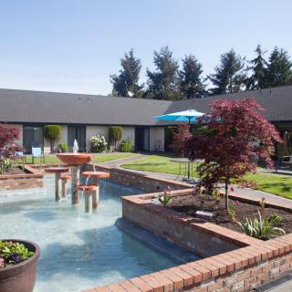 A serene courtyard featuring a central fountain surrounded by lush greenery and brick borders.