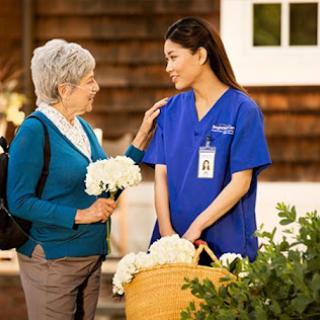 A caring nurse shares a moment with an elderly woman, surrounded by flowers in a warm outdoor setting.