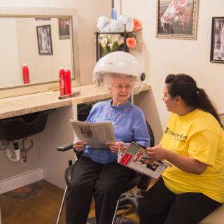 An elderly woman sits under a hair dryer while reading a magazine, chatting with a hairdresser in a salon.
