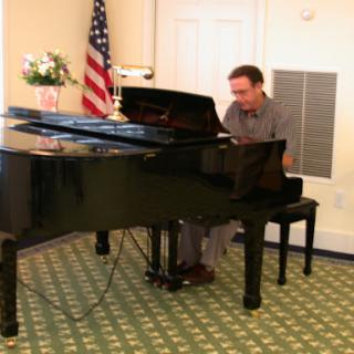 A musician performing on a black grand piano in an indoor setting, accompanied by an American flag.