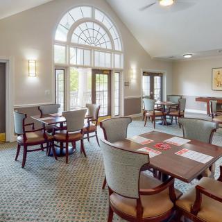 A well-lit dining room with several tables and chairs arranged around a carpeted floor, featuring large arched windows.