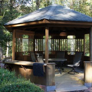 A wooden gazebo surrounded by greenery, featuring outdoor furniture.