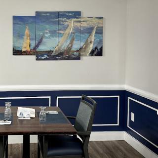 A cozy dining area featuring a wooden table, comfortable chairs, and a vibrant painting of sailboats on a blue wall.