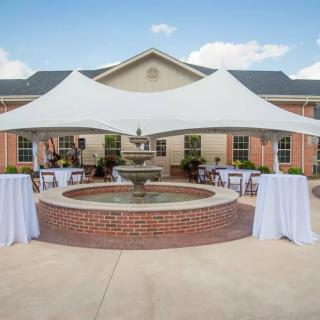 An outdoor event setup featuring a large tent, fountain, and several tables draped with white cloths in front of a brick building.
