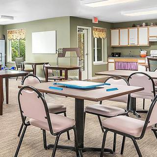 A well-lit multipurpose room featuring tables and chairs, resembling a classroom or community space with a kitchen area in the background.