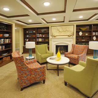 A cozy library reading room featuring armchairs, a coffee table, and bookshelves surrounding a fireplace.