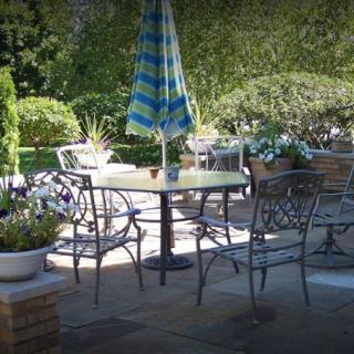 A tranquil patio setting with a table, chairs, and a sun umbrella surrounded by lush greenery.