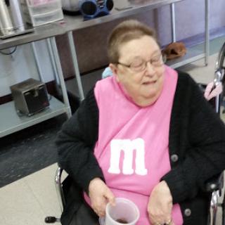 An elderly woman in a pink shirt sits comfortably in her wheelchair, smiling while holding a cup.