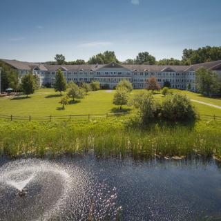 A scenic view of a large building surrounded by greenery with a pond and fountain in the foreground.