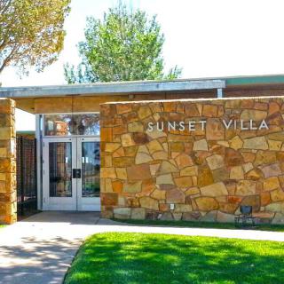 The entrance of a building with a stone facade and 'SUNSET VILLA' signage, surrounded by grass and trees.