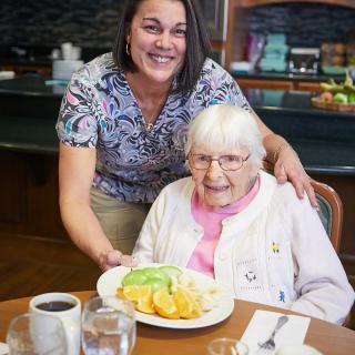 A caregiver smiles as she presents a meal to an elderly woman at the dining table.