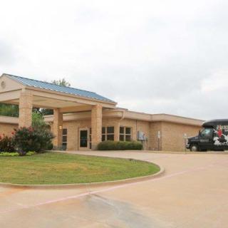 The image shows the entrance of a hospital with a driveway and vehicles parked nearby.