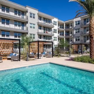 A modern apartment complex with a swimming pool surrounded by outdoor seating and palm trees.