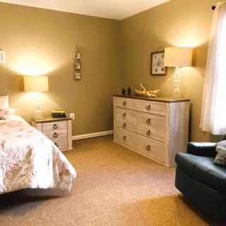 A cozy bedroom with warm lighting, featuring a bed, dresser, and armchair.
